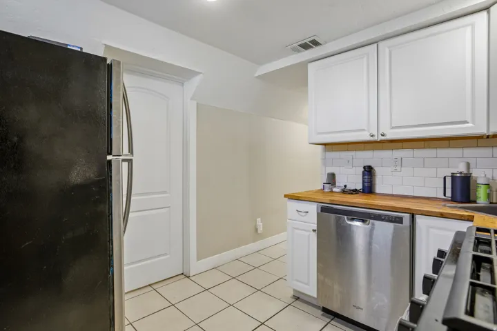 White shaker-style cabinetry, wood-finish countertops, stainless steel dishwasher, white subway tile backsplash, and tile flooring