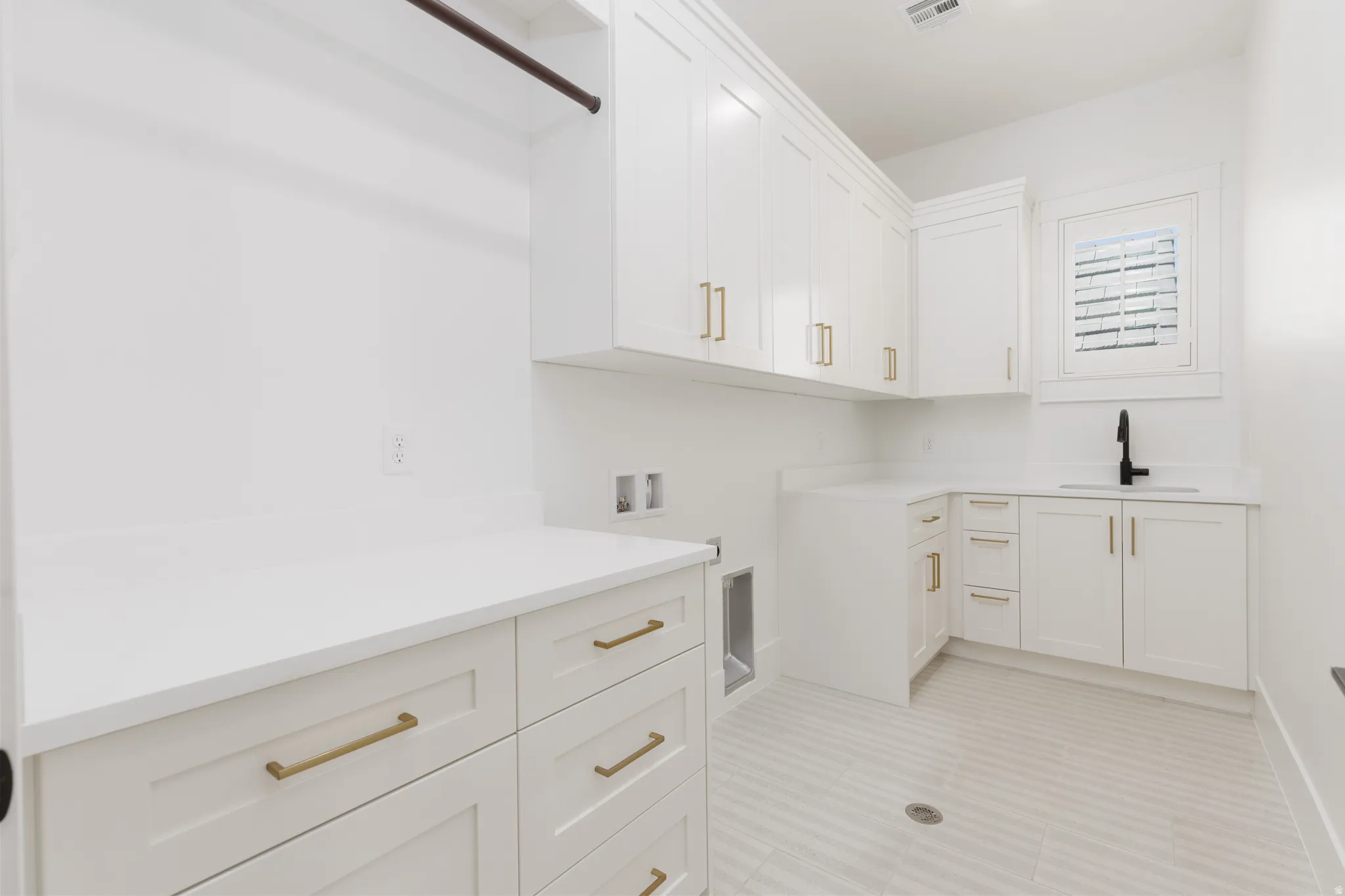 White cabinetry with gold-finish hardware, light-toned countertops, a stainless-steel sink with a matte black faucet, and a window with horizontal blinds