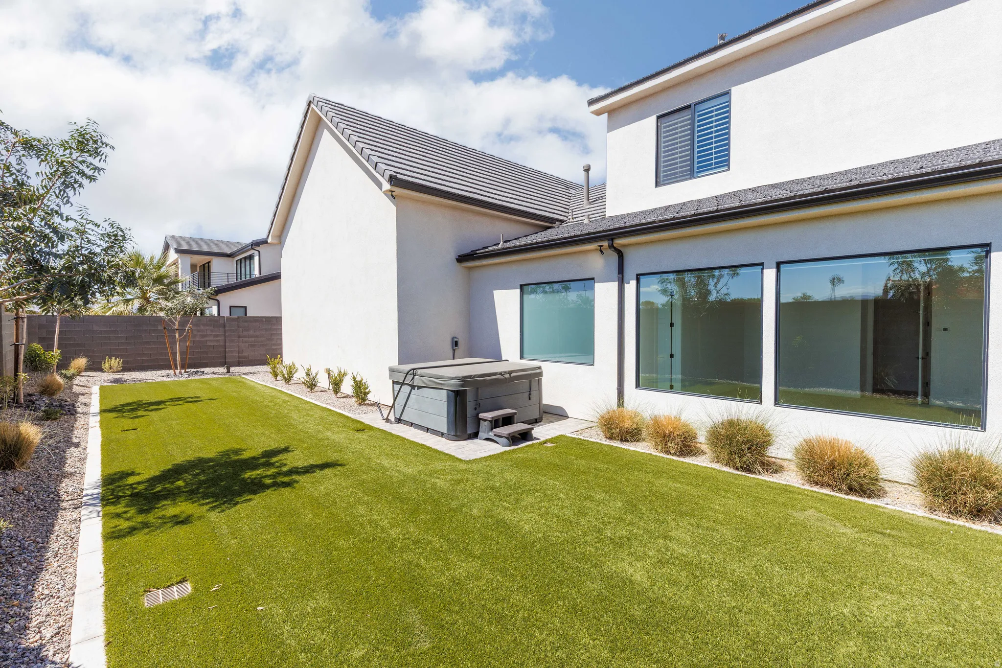 Expansive backyard featuring artificial turf, a hot tub, full-height windows, a gravel border, and a concrete patio