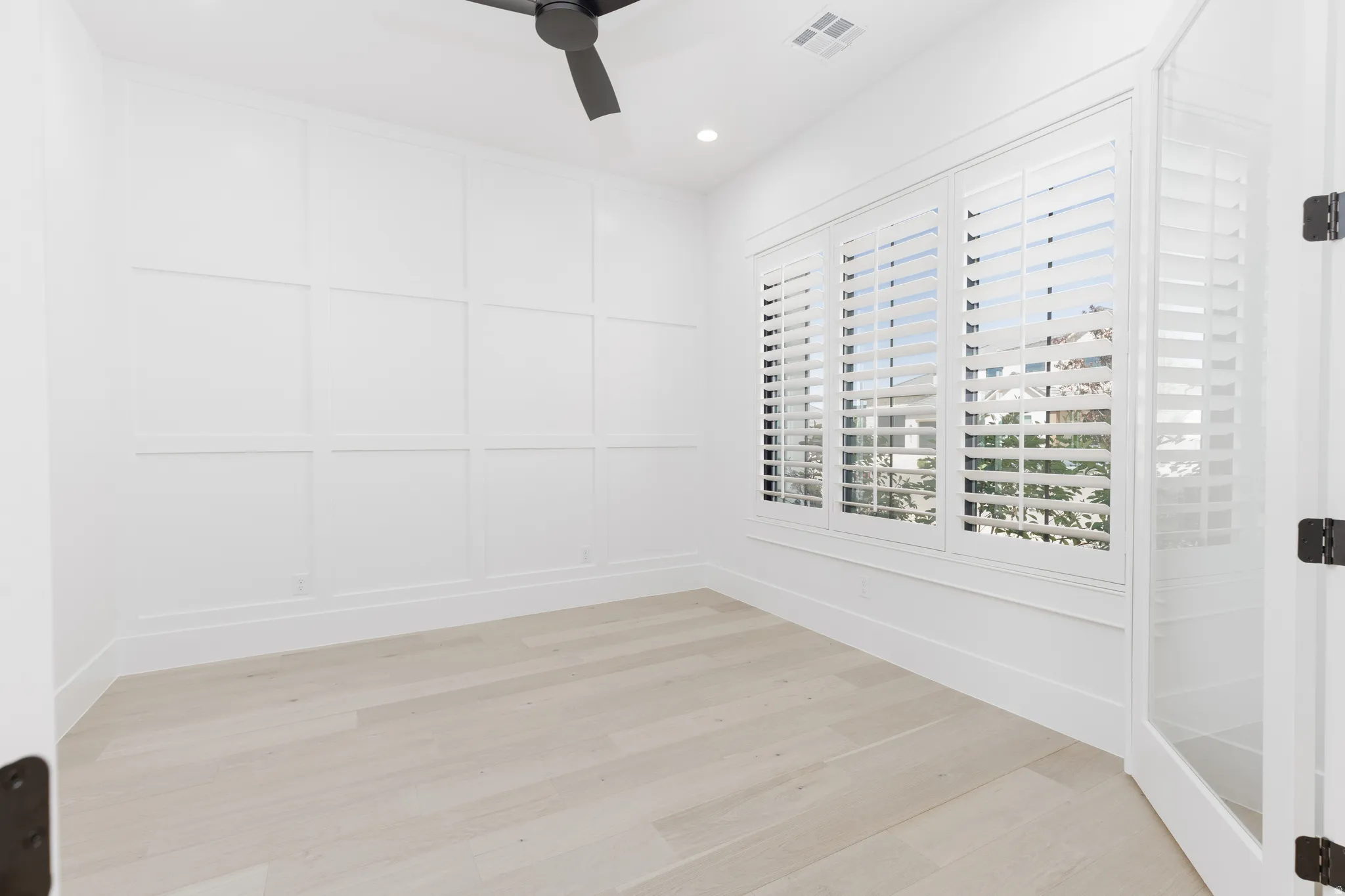 White paneled accent wall, wood-finish flooring, multiple windows with white plantation shutters, glass-paneled door, and a black ceiling fan