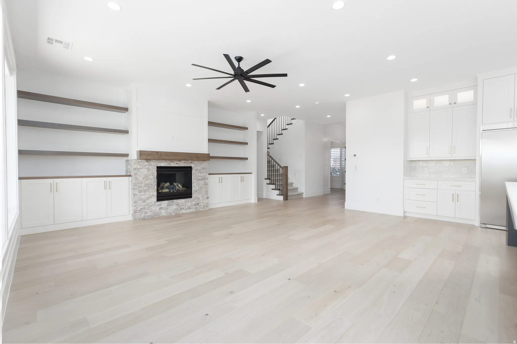 Spacious living area featuring light wood-finish flooring, a stone-faced fireplace with a wood mantle, built-in shelving and cabinetry, and a ceiling fan