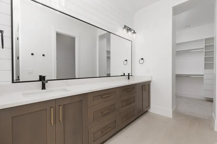 Dual vanity with integrated sinks, expansive mirror, wood-finish cabinetry with brass hardware, and tile flooring