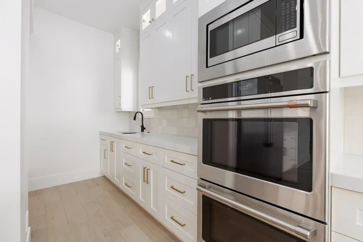 Built-in stainless steel appliances, white cabinetry with gold-tone hardware, white countertop, white tile backsplash, and wood-finish flooring