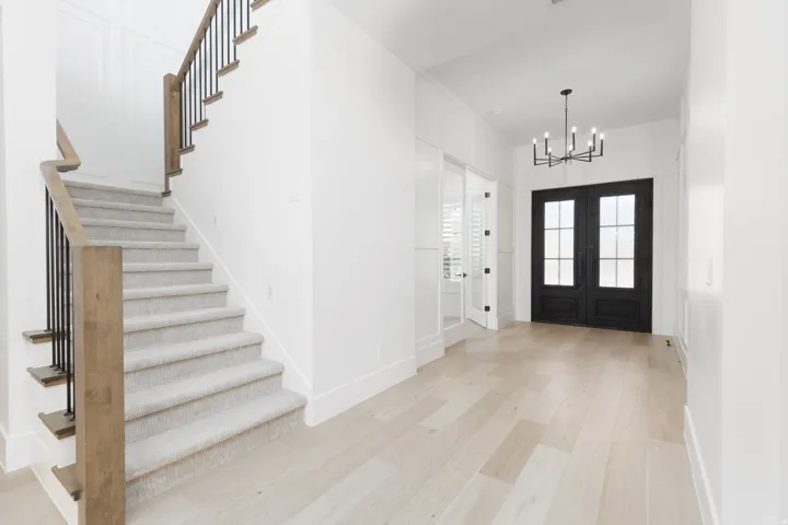 Light wood-finish flooring, a carpeted staircase with wood and metal railings, and a black double-door entry with glass panels