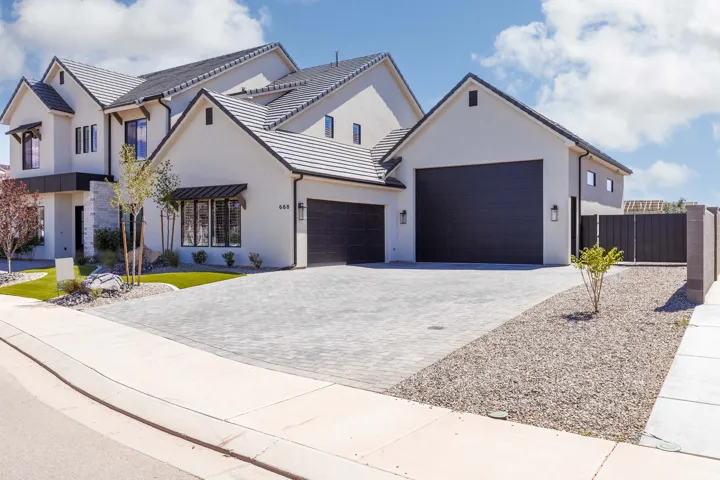 Modern stucco exterior with a multi-gabled roofline, two dark-toned garage doors, a paved driveway, and mature landscaping