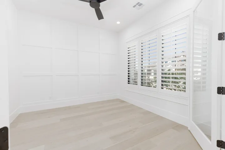 White paneled accent wall, wood-finish flooring, multiple windows with white plantation shutters, glass-paneled door, and a black ceiling fan