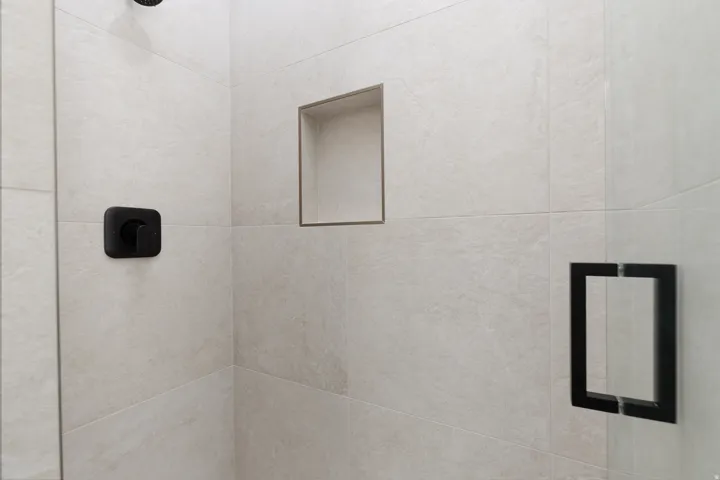Tiled shower with a recessed niche, black square shower controls, and a frameless glass shower enclosure with a black handle