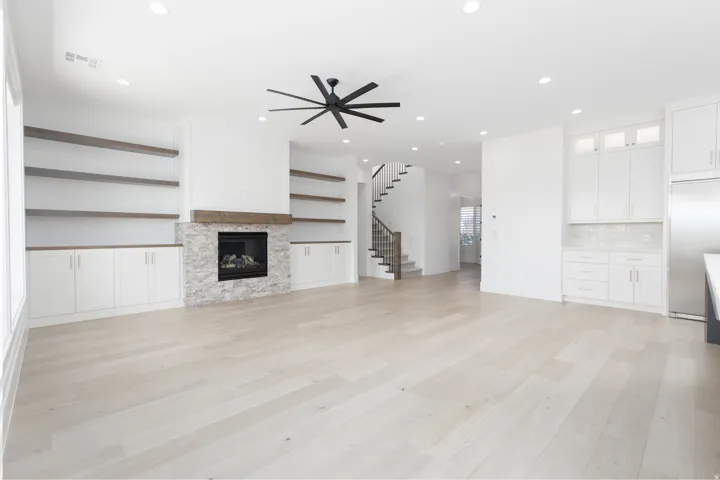 Spacious living area featuring light wood-finish flooring, a stone-faced fireplace with a wood mantle, built-in shelving and cabinetry, and a ceiling fan