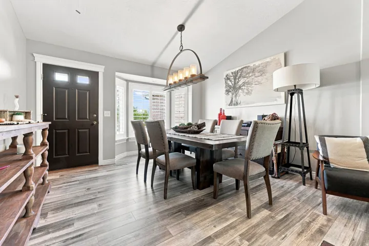 Open concept wood-finish flooring, bay window with plantation shutters, vaulted ceiling, and a dark paneled entry door with transom windows