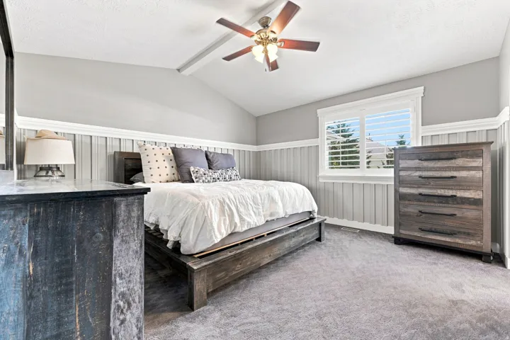 Vaulted ceiling with exposed beam, ceiling fan with light fixture, carpeted flooring, window with plantation shutters, and vertical paneling with crown molding