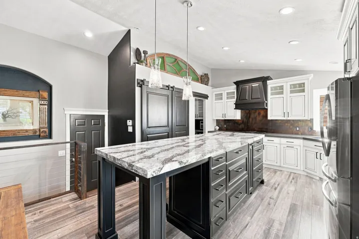 Spacious kitchen featuring a large island with a patterned countertop, white perimeter cabinetry, a dark wood-finish range hood, and wood-finish flooring