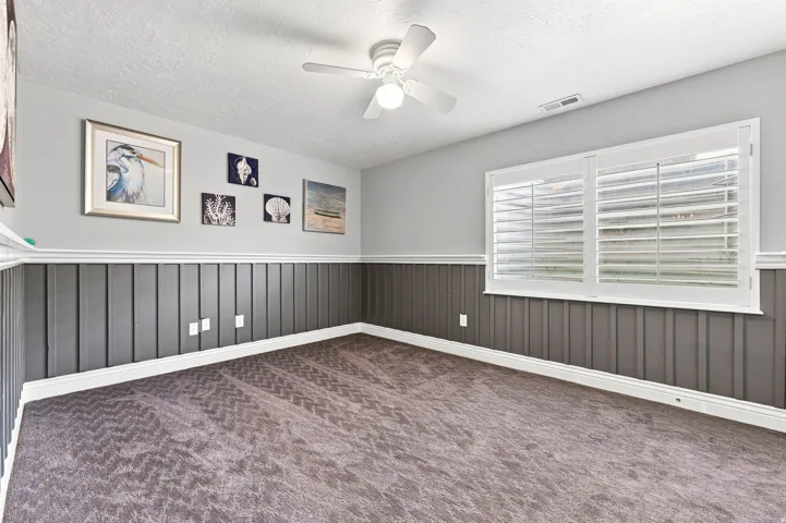 Board and batten wainscoting, textured carpet, ceiling fan, and window with plantation shutters