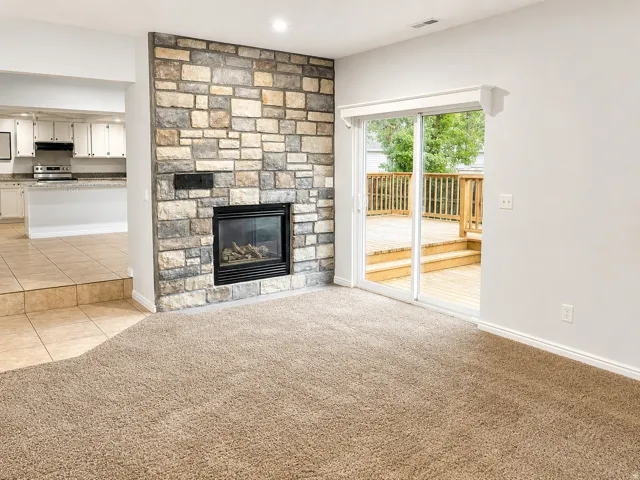 Stone-faced fireplace with a gas insert, sliding glass door to an outdoor wood deck, and a kitchen with white cabinetry and light-toned countertops