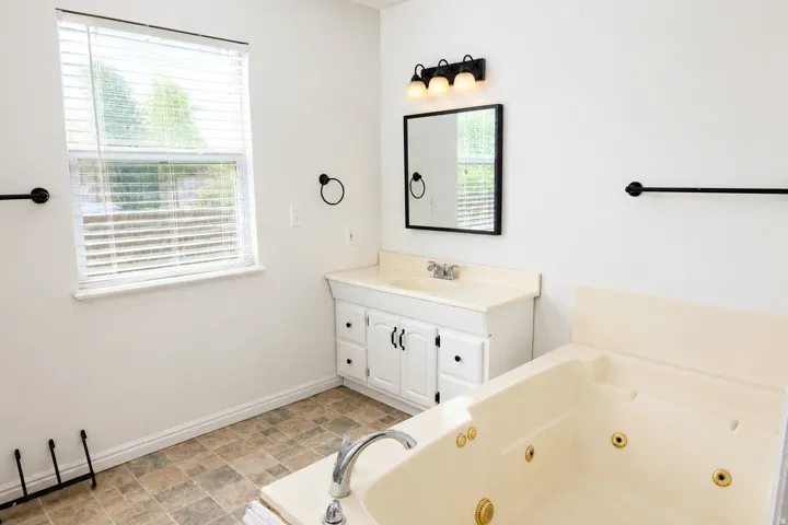 Spa-inspired primary bathroom featuring a soaking tub with integrated jets, dual vanity with white cabinetry and brushed nickel hardware, ceramic tile flooring, framed mirror with sconce lighting, and large window with privacy shutters