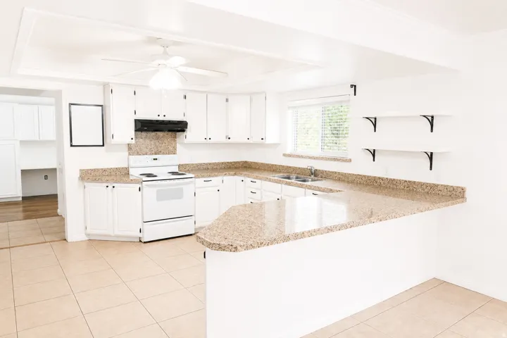 Kitchen featuring white cabinetry, light-toned stone countertops, a dual basin stainless steel sink, a white range with overhead exhaust, and ceramic tile flooring