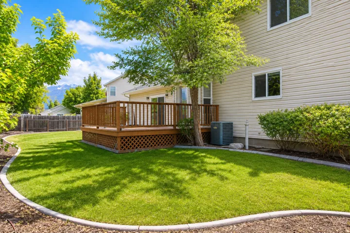 Expansive backyard featuring a large wooden deck with lattice skirting, a manicured lawn, concrete landscape curbing, and mature trees