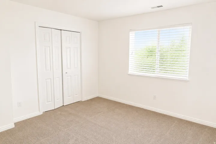Bright, light-filled bedroom featuring neutral carpeting, a large window with horizontal blinds providing natural light, and a spacious closet with bi-fold doors