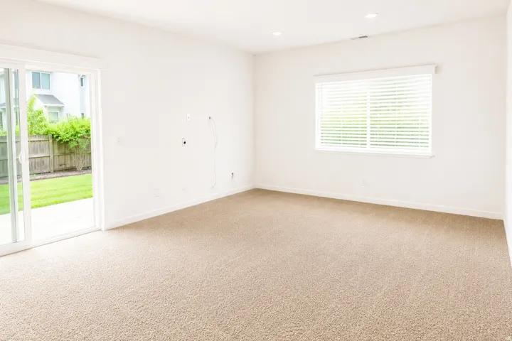 Recessed ceiling lighting, a large window with blinds, soft neutral carpeting, and a sliding glass door opening to a concrete patio and fenced yard