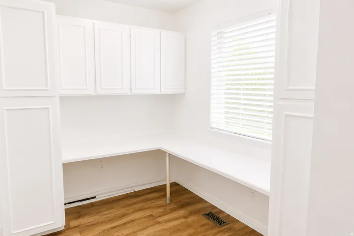Built-in desk with extensive white cabinetry, durable wood-finish flooring, and natural light from a window with blinds