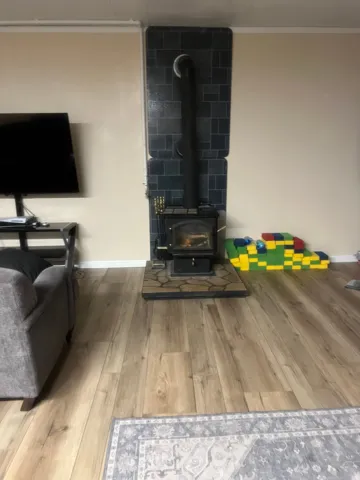 Spacious living area featuring warm wood-finish flooring, a traditional wood-burning stove with dark tile surround and chimney breast, and ample natural light from large windows