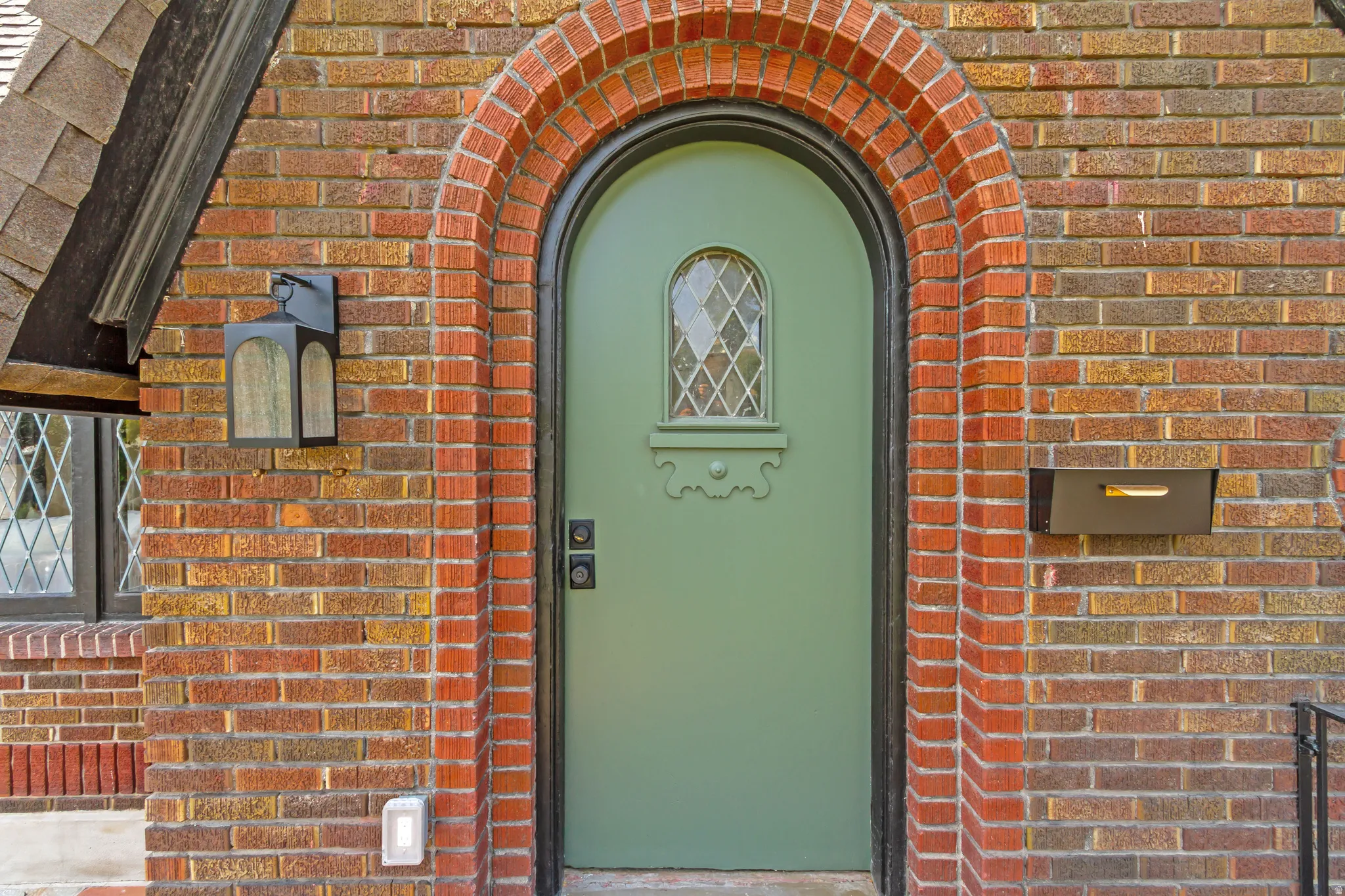Brick exterior with an arched entry, featuring a sage green door with a diamond-pane window, an exterior lantern fixture, and a built-in mailbox