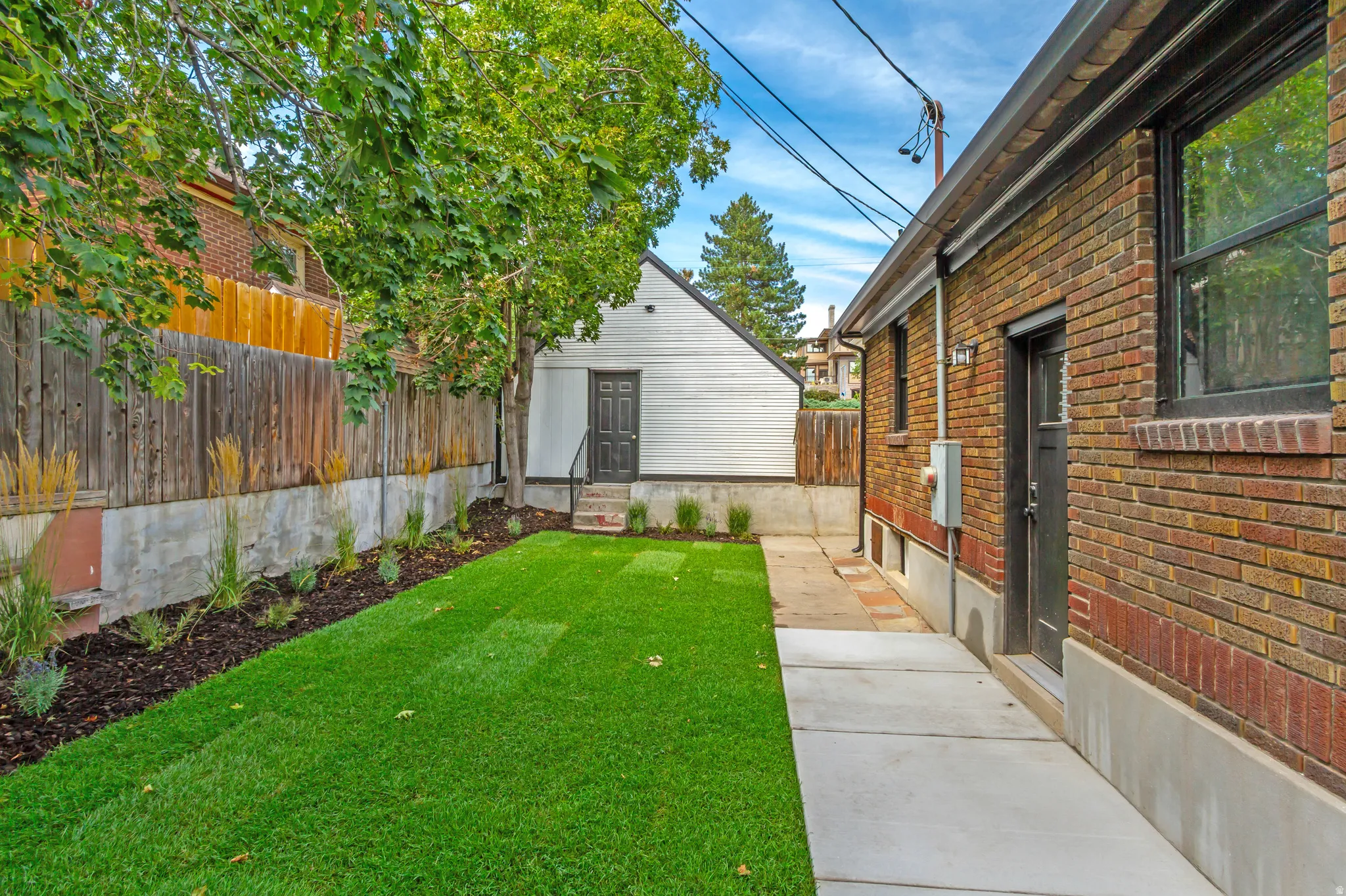 Manicured lawn with mature trees, brick exterior, concrete walkway, detached structure, and privacy fencing