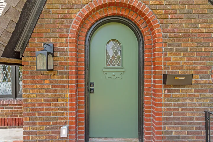 Brick exterior with an arched entry, featuring a sage green door with a diamond-pane window, an exterior lantern fixture, and a built-in mailbox