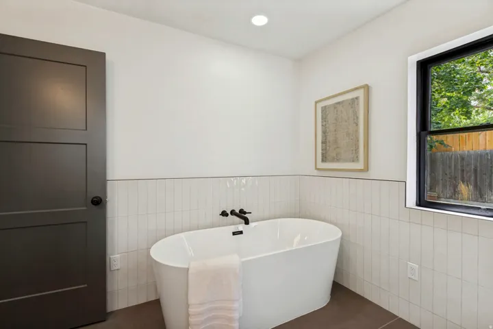 Freestanding soaking tub with matte black fixtures, vertical subway tile wainscoting, a dark frame window, and a recessed ceiling light