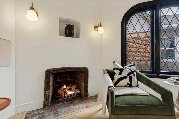 Cozy sitting room featuring a traditional cast-iron fireplace with decorative tilework surround, large leaded-glass windows with black metal framing, brass pendant lighting fixtures, and upholstered seating with throw pillows and blankets