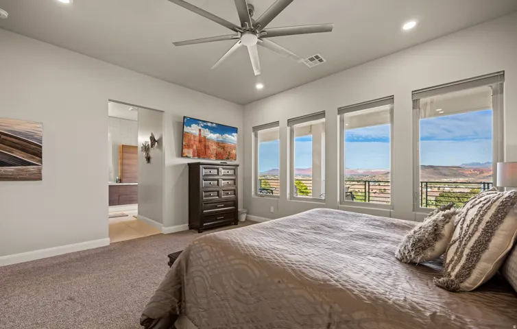 Spacious bedroom with plush carpeting, recessed lighting, a modern ceiling fan, and multiple floor-to-ceiling windows with roller shades