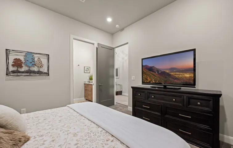 Master bedroom with recessed ceiling lighting, ensuite bathroom access, black wood-finish dresser with multiple drawers, flat-screen television mounted on wall, and large window with frosted glass shower enclosure visible through open doorway
