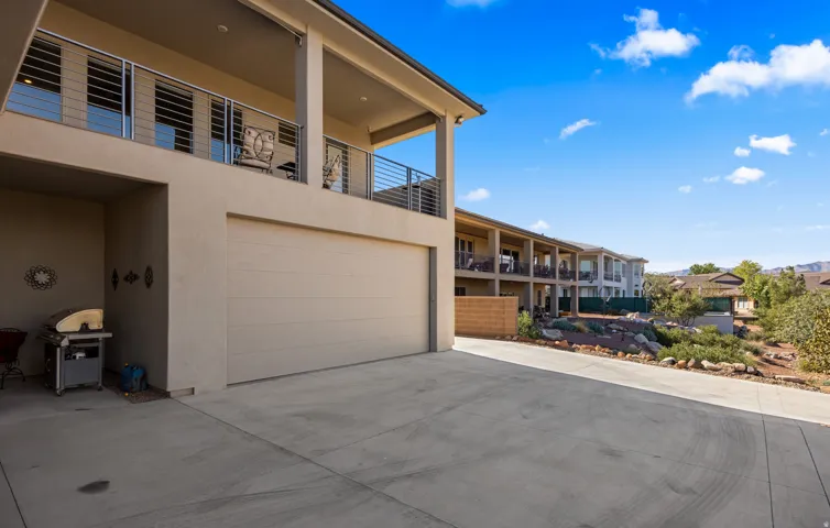 Modern home featuring a spacious concrete driveway, three-car garage with paneled door, horizontal metal railing on the upper-level balcony, and clean-lined architectural design with stucco exterior and large glass sliding doors opening to covered outdoor