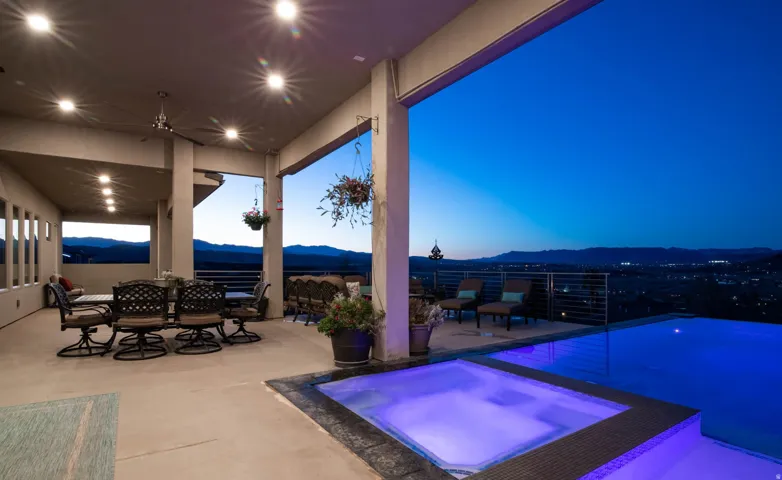 Sophisticated outdoor entertaining space featuring a resort-style illuminated spa with integrated water features, expansive covered loggia with recessed lighting and structural columns, polished concrete flooring, and panoramic landscape vistas extending