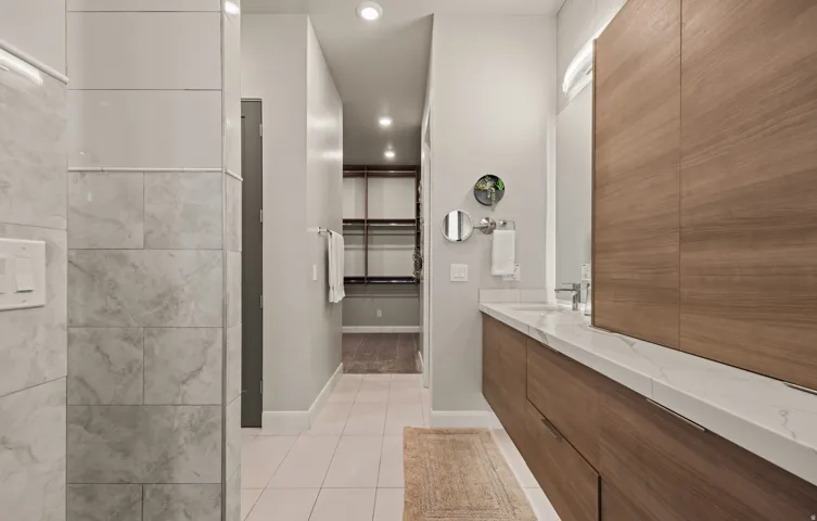 Spa-inspired primary bathroom featuring a spacious walk-in shower with large-format tile surround, dual vanity with white quartz countertop, warm wood-finish cabinetry, polished chrome fixtures, and recessed lighting throughout