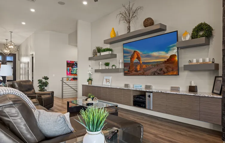 Modern open-concept living area featuring a wood-finish plank floor, gray horizontal panel cabinetry with integrated storage, floating shelves with decorative display space, a wall-mounted flat-screen television, and recessed ceiling lighting