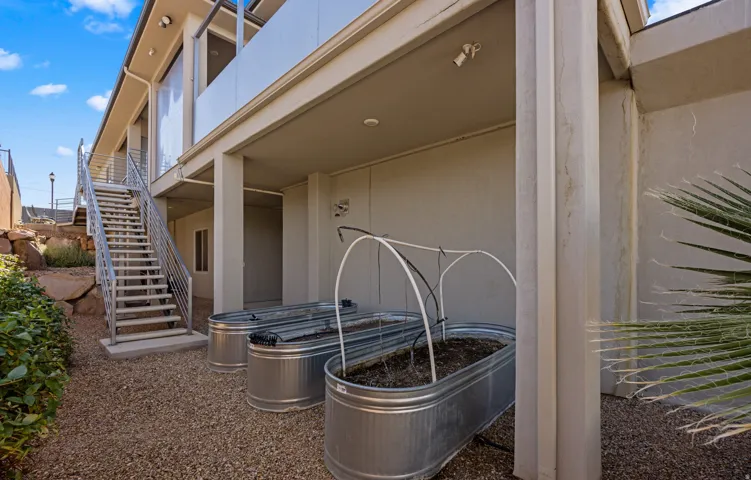 Ground-level patio with gravel landscaping, featuring three galvanized metal planters, a modern exterior staircase with metal railings, and a covered patio area with support columns