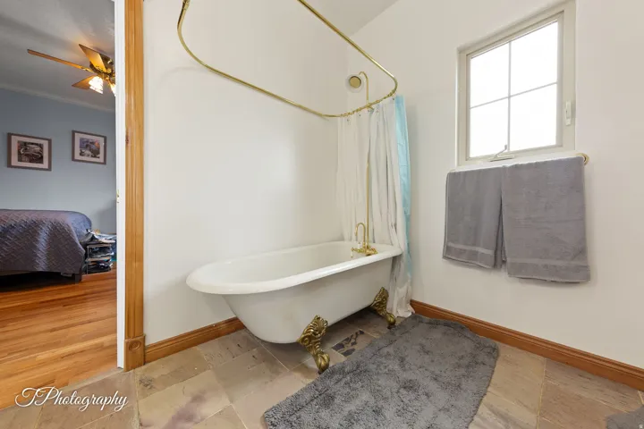 Clawfoot tub with brass fixtures, natural stone tile flooring, and a single-pane window