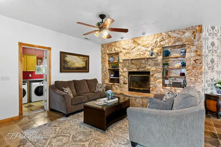 Welcoming living room showcasing a wood-burning fireplace with stone surround and built-in shelving, ceiling fan with lighting, hardwood flooring, and direct access to the laundry area
