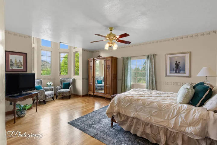 Spacious primary bedroom suite featuring hardwood flooring, multiple windows with natural light, ceiling fan with brass finish, and an adjoining sitting area with seating capacity