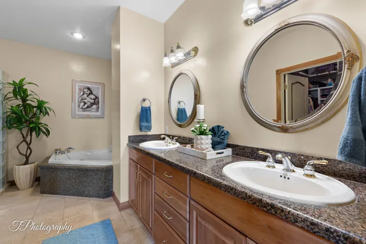 Spa-inspired primary bathroom featuring dual vanities with stone countertops, two oval mirrors with integrated lighting, and a soaking tub