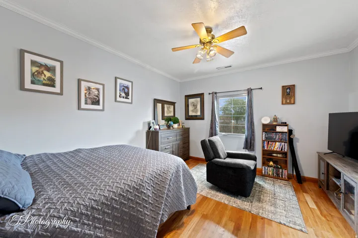 Light gray walls with crown molding, wood-finish flooring, a ceiling fan with integrated lighting, and a single window with dark-toned curtains