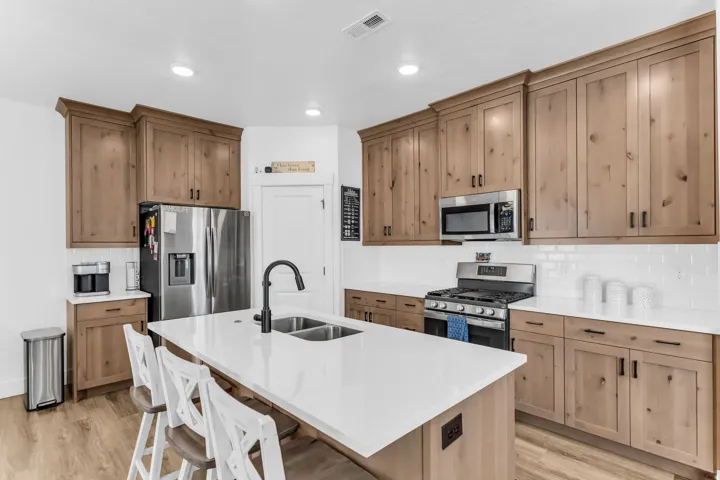Kitchen featuring natural wood-finish cabinetry, white subway tile backsplash, stainless steel appliances, a center island with a double basin sink, and wood-finish flooring