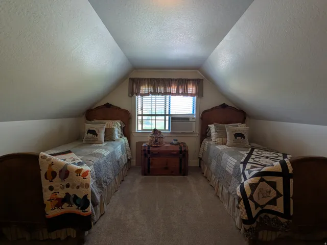 Charming attic bedroom featuring dual twin beds with wood headboards, sloped ceilings that follow the roofline, and a dormer window with natural light and blue-paned glass detailing