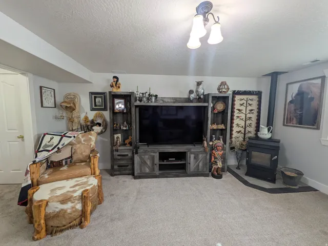 Recessed ceiling, neutral carpet, wood-finish media console with shelving, wood-burning stove with hearth, and wall-mounted lighting fixture