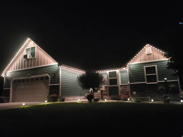Gable roofline with vertical siding accents, horizontal siding facade, stone veneer detailing, attached garage with panel door, and exterior illumination