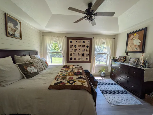 Master Bedroom with on suite and Vaulted ceiling, ceiling fan with light fixture, wood-finish flooring, two windows with white curtains, horizontal wall paneling