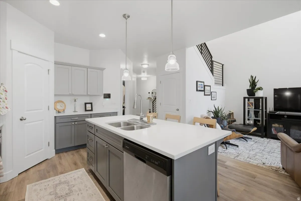 Kitchen featuring light gray cabinetry, a white countertop island with dual sinks, stainless steel dishwasher, and wood-finish flooring