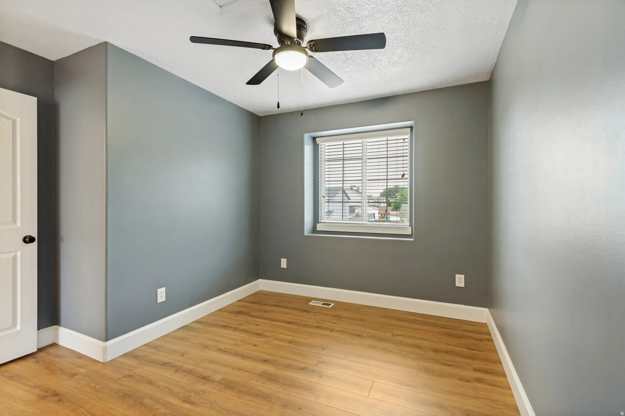 Neutral gray walls, wood-finish flooring, white window blinds, white interior door with black hardware, and a ceiling fan with integrated lighting