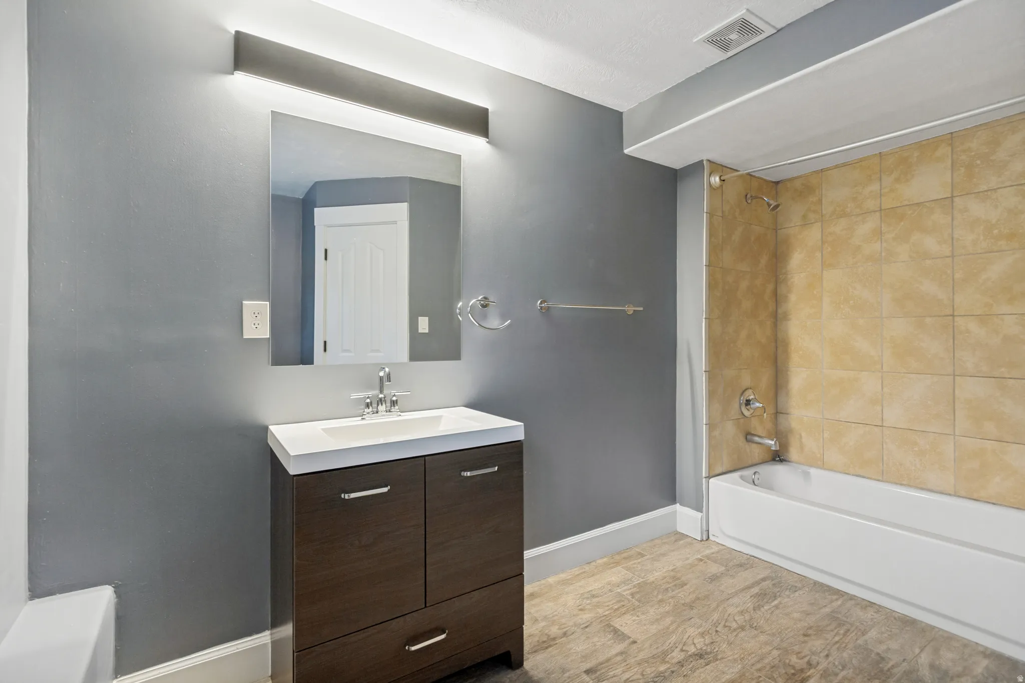 Bathroom featuring a dark wood-finish vanity with an integrated white sink, a vanity mirror with overhead lighting, a bathtub with tiled surround, and wood-finish flooring