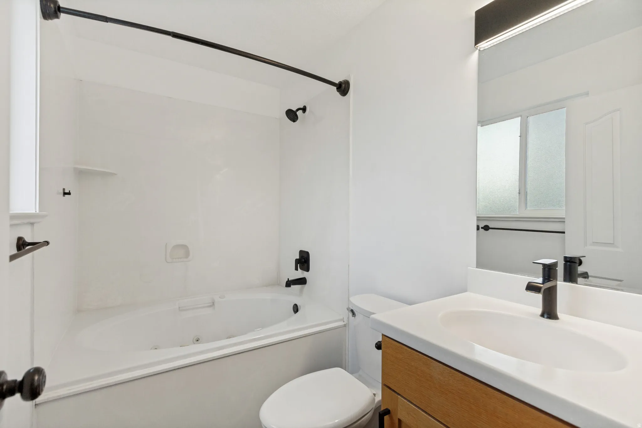 Bathroom featuring a jetted tub, a vanity with a light wood-finish cabinet, a white countertop with an integrated sink, and matte black fixtures